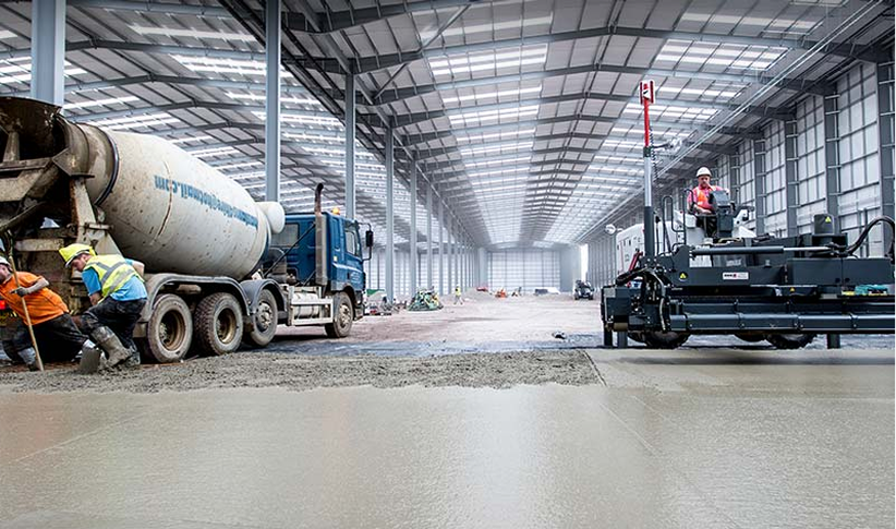 Workers operating construction equipment and pouring concrete inside a large industrial warehouse under construction.