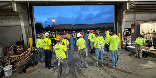 Group of construction workers wearing safety vests and hard hats gathered inside an industrial warehouse during an early morning safety briefing.